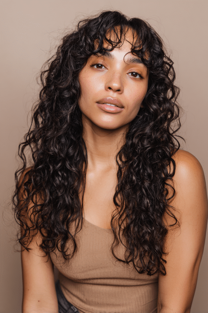 A vibrant American female model in her 20s with chest-length curly Hime Cut long hair featuring bouncy curls, defined sidelocks, and soft bangs, styled vibrantly, photographed against a seamless soft taupe backdrop with a matte finish. She wears subtle makeup, a trendy outfit, and has a warm skin tone with a friendly expression, captured with soft natural lighting.