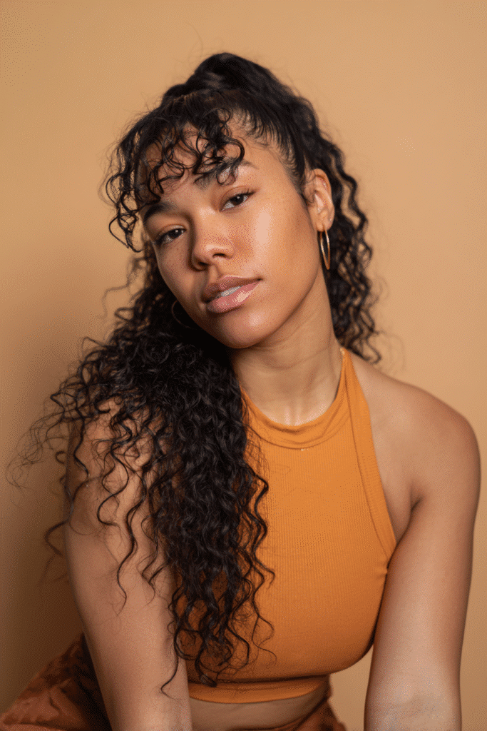 A vibrant Black female model in her 20s with shoulder-to-waist-length curly wig featuring a lace frontal, styled in a half-up high ponytail with voluminous curls flowing down, photographed against a seamless warm caramel backdrop with a matte finish. She has subtle makeup, a trendy outfit, a warm skin tone, and a friendly expression, captured with soft natural lighting.