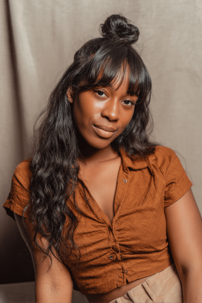 A vibrant Black female model in her 20s with shoulder-to-waist-length deep wave wig, styled in a half-up loose top knot with deep wave strands flowing down, photographed against a seamless soft taupe backdrop with a smooth texture. She has subtle makeup, a trendy outfit, a warm skin tone, and a friendly expression, captured with soft natural lighting.