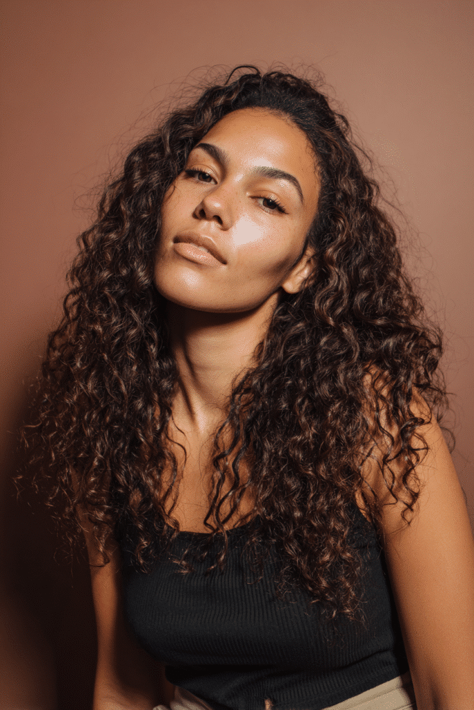 A vibrant female model in her 20s with shoulder-to-waist-length curly half wig, styled with defined curls for non-braiders and tight, springy curls flowing down, photographed against a seamless soft plum backdrop with a velvety texture. She wears subtle makeup, a trendy outfit, and has a warm skin tone with a friendly expression, captured with soft natural lighting.