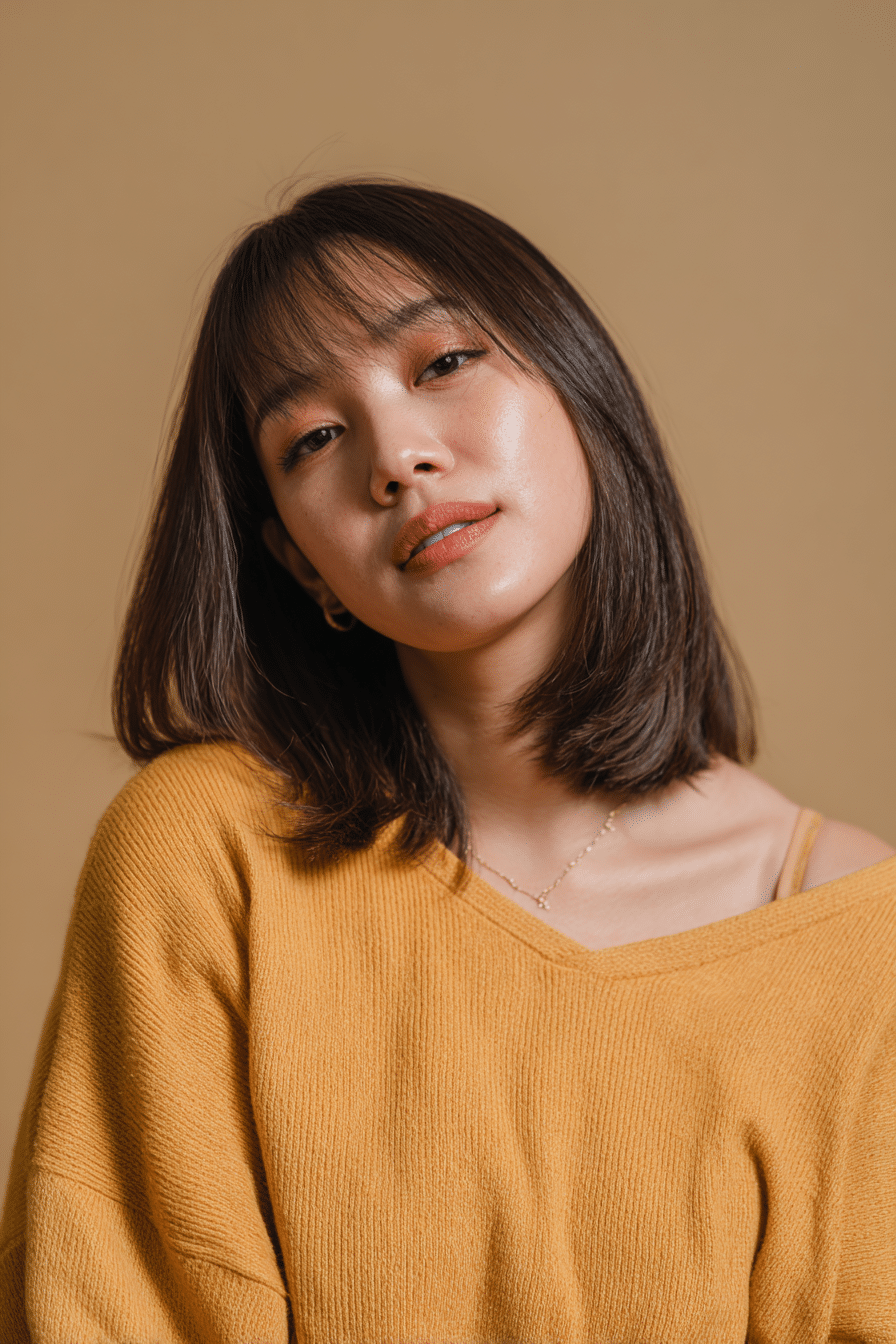 A vibrant American female model in her 20s with chest-length straight Hime Cut long hair featuring prominently defined double sidelocks and straight ends with no bangs, styled elegantly, photographed against a seamless warm beige backdrop with a satin finish. She has subtle makeup, a trendy outfit, a warm skin tone, and a friendly expression, captured with soft natural lighting.
