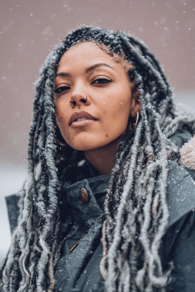A vibrant female model in her 20s with shoulder-to-waist-length faux locs, styled with a protective and natural flow, captured in a close-up shot focusing on the hair, photographed against a seamless soft mauve backdrop with a glossy sheen. She has subtle makeup, a stylish winter coat, a warm skin tone, and a confident expression, captured with cozy winter lighting and soft snowflakes.