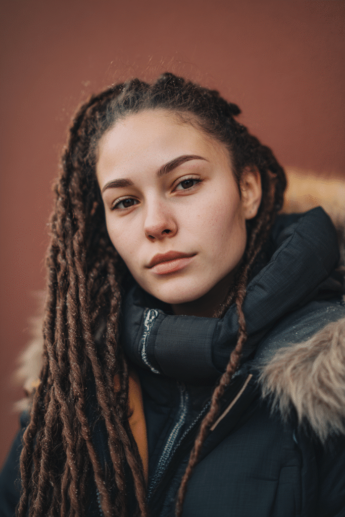 A vibrant female model in her 20s with shoulder-to-waist-length flat twists, styled with a protective and sleek design, captured in a close-up shot focusing on the hair, photographed against a seamless warm plum backdrop with a satin finish. She has subtle makeup, a stylish winter coat, a warm skin tone, and a confident expression, captured with cozy winter lighting and a frosty glow.