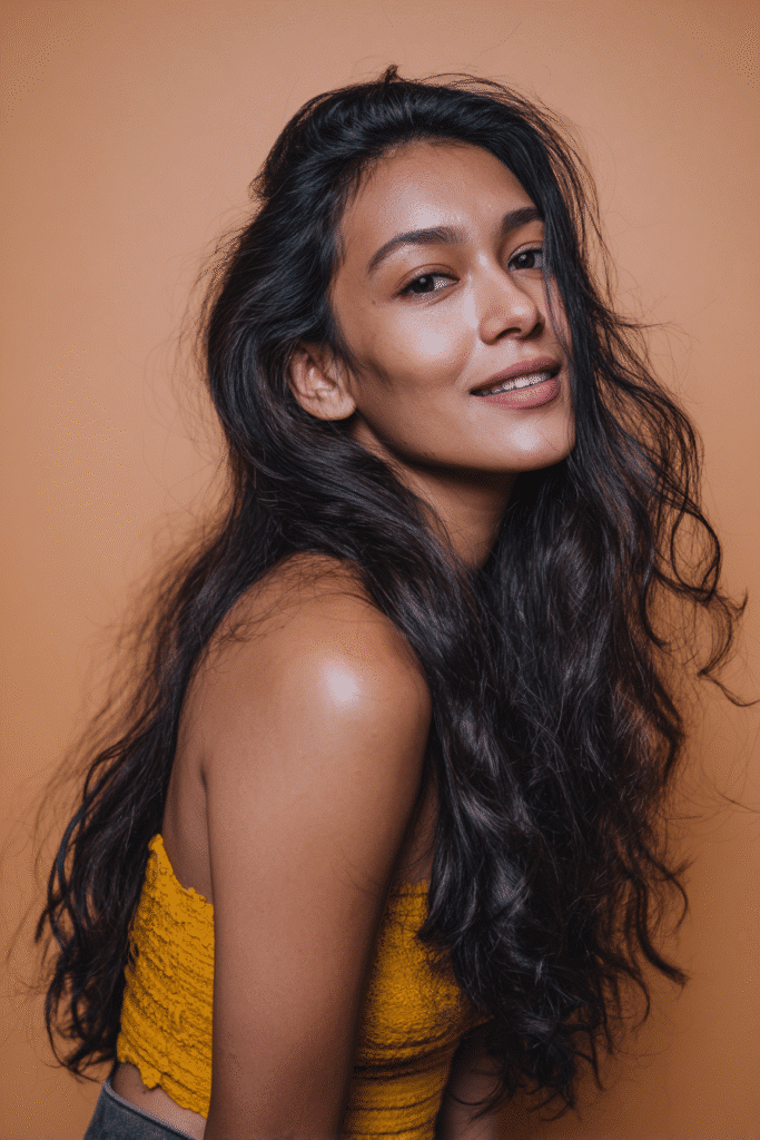 A vibrant female model in her 20s with shoulder-to-waist-length wavy half wig, styled with a voluminous flip over and loose waves flowing down, photographed against a seamless warm coral backdrop with a matte finish. She has subtle makeup, a trendy outfit, a warm skin tone, and a friendly expression, captured with soft natural lighting.