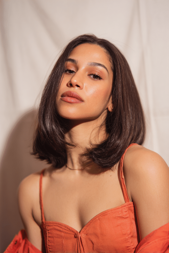 A vibrant female model in her 20s with shoulder-to-waist-length straight hair, styled in a frontal baddie wig with styled baby hair along the hairline, framed from chest-level upward, photographed against a seamless soft ivory backdrop with a glossy sheen. She has bold makeup, a trendy outfit, a warm skin tone, and a confident expression, captured with soft natural lighting.