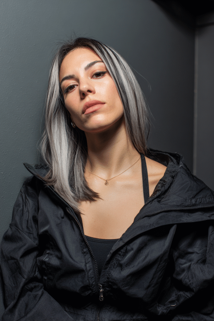 A vibrant female model in her 20s with shoulder-to-waist-length straight hair featuring gray highlights, styled in a sleek straight baddie wig, framed from chest-level upward, photographed against a seamless soft charcoal backdrop with a silky finish. She has bold makeup, a trendy outfit, a warm skin tone, and a confident expression, captured with soft natural lighting.