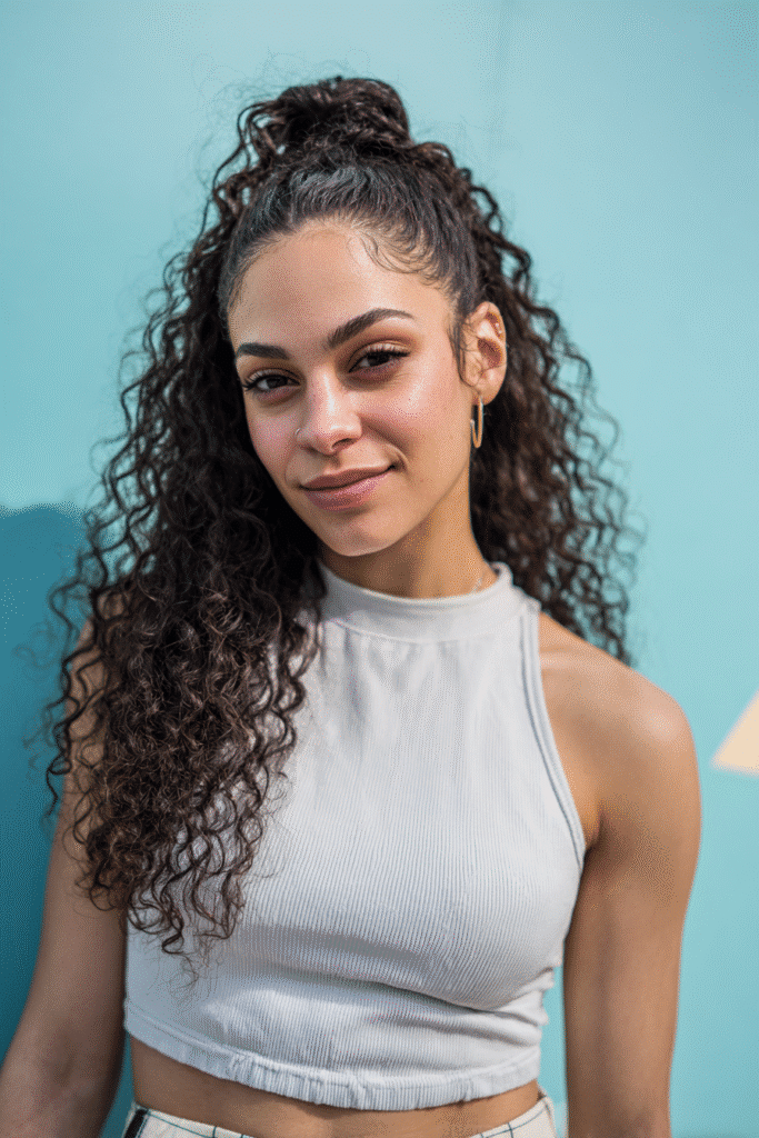 A vibrant female model in her 20s with shoulder-to-waist-length curly half wig, styled with a small twisted bun half up and curls flowing down, photographed against a seamless light aqua backdrop with a smooth surface. She wears subtle makeup, a trendy outfit, and has a warm skin tone with a friendly expression, captured with soft natural lighting.