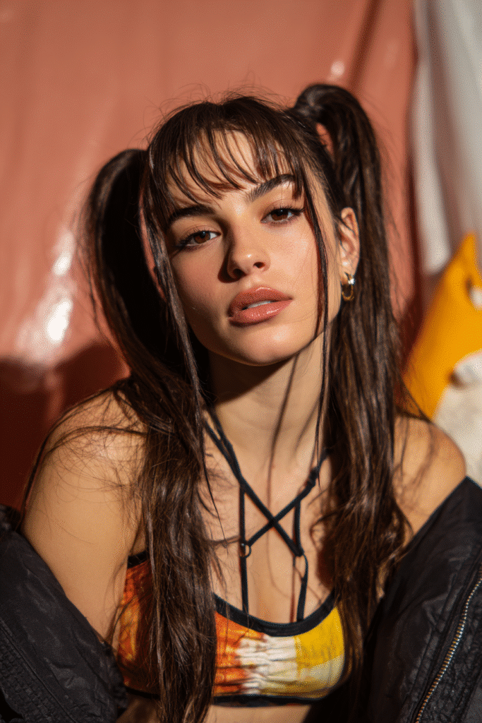 A vibrant female model in her 20s with shoulder-to-waist-length straight hair, styled in a half-up pigtail baddie wig with two high ponytails, framed from chest-level upward, photographed against a seamless warm rose backdrop with a glossy sheen. She has bold makeup, a trendy outfit, a warm skin tone, and a confident expression, captured with soft natural lighting.