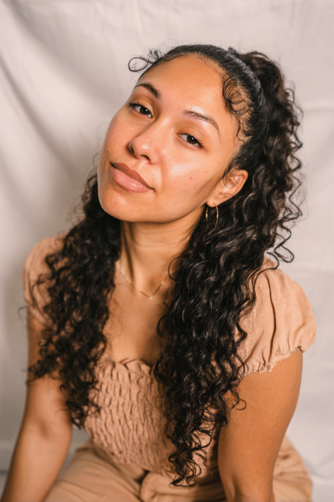 A vibrant Black female model in her 20s with shoulder-to-waist-length curly wig, styled in a half-updo with cute, tight curls flowing down, photographed against a seamless soft ivory backdrop with a satin finish. She has subtle makeup, a trendy outfit, a warm skin tone, and a friendly expression, captured with soft natural lighting.
