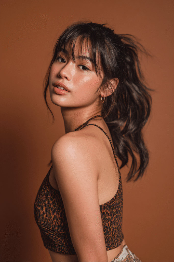 A vibrant female model in her 20s with shoulder-to-waist-length straight half wig, styled with a Fauk Hawk featuring intricate twists and volume at the crown, with soft, loose strands cascading naturally down the sides and back, photographed against a seamless warm bronze backdrop with a matte finish. She wears subtle makeup, a instagram trendy outfit, and has a warm skin tone with a friendly expression, captured with soft natural lighting.