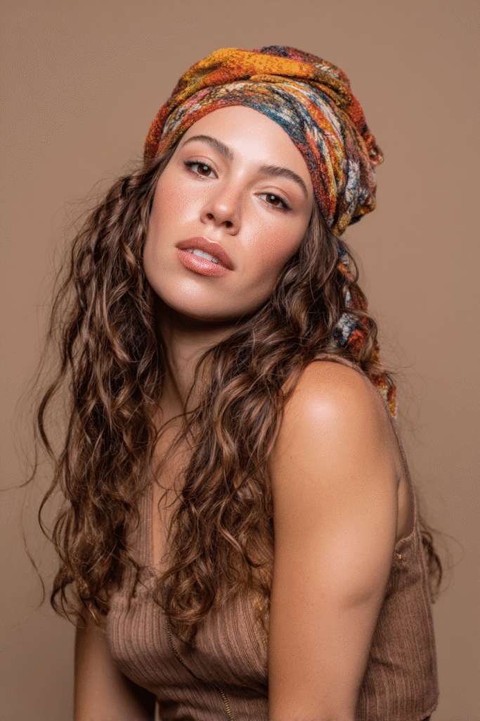 A vibrant female model in her 20s with shoulder-to-waist-length wavy half wig, styled with a colorful scarf woven into the hair and loose waves flowing down, photographed against a seamless warm taupe backdrop with a matte finish. She has subtle makeup, a trendy outfit, a warm skin tone, and a friendly expression, captured with soft natural lighting.