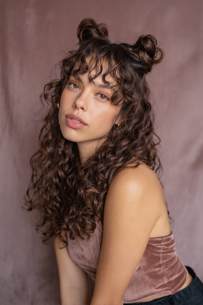 A vibrant female model in her 20s with shoulder-to-waist-length curly half wig, styled with space buns and loose curls flowing down, photographed against a seamless soft mauve backdrop with a velvety texture. She wears subtle makeup, a trendy outfit, and has a warm skin tone with a friendly expression, captured with soft natural lighting.