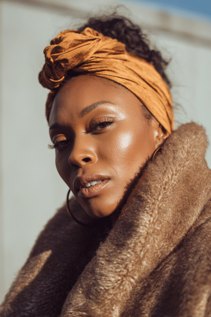 A vibrant young woman in her 20s wearing a stylish winter coat, showcasing an elegant headwrap as a protective winter hairstyle. The close-up portrait focuses on the detailed headwrap and covered hair, enhanced by cozy winter lighting with a frosty glow. She has subtle makeup, a warm natural skin tone, and a confident expression, posing against a seamless warm gold backdrop with a glossy sheen, captured in high-quality Instagram-style photography.