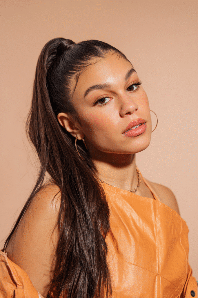 A vibrant female model in her 20s with shoulder-to-waist-length straight half wig, styled in a high ponytail with sleek strands flowing down, photographed against a seamless soft peach backdrop with a satin finish. She has subtle makeup, a trendy outfit, a warm skin tone, and a friendly expression, captured with soft natural lighting.