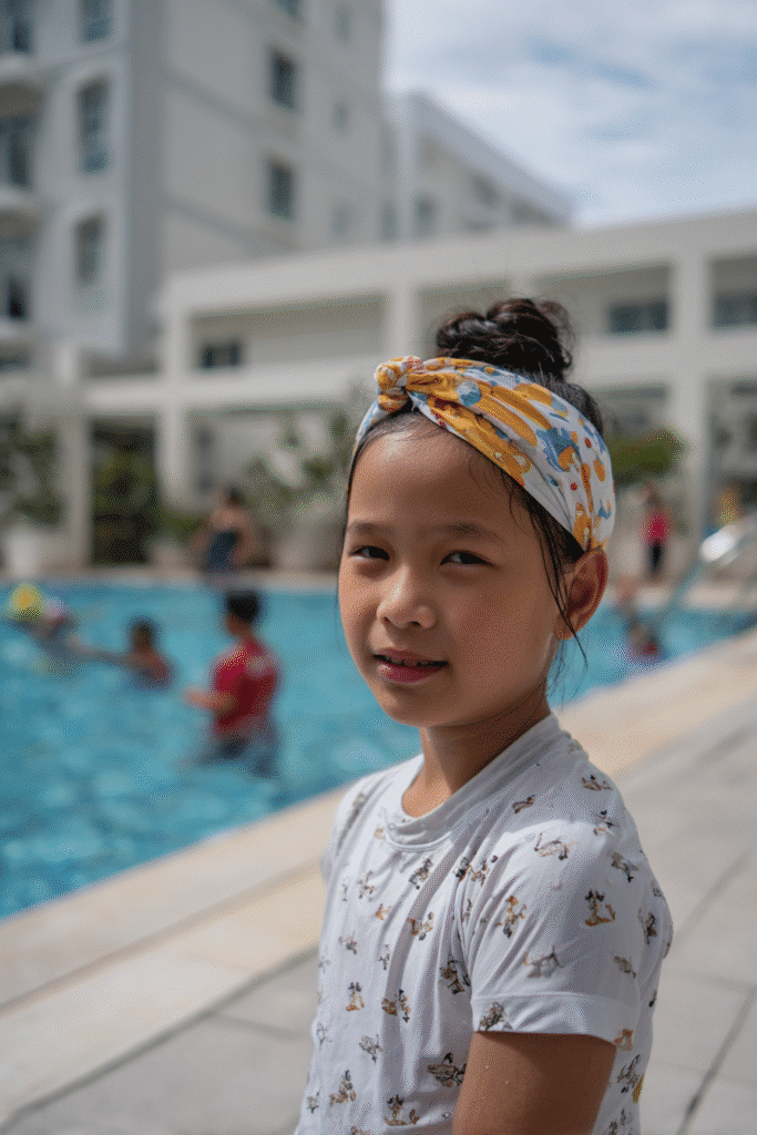 American teenage girl with a curly high puff secured with a colorful headband, modest swim shirt, standing near a lively pool area.