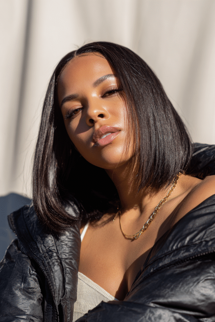 A vibrant female model in her 20s with shoulder-to-waist-length straight hair in jet black, styled in a quick weave baddie wig with a sleek and bold finish, framed from chest-level upward, photographed against a seamless light ivory backdrop with a glossy finish. She has bold makeup, a trendy outfit, a warm skin tone, and a confident expression, captured with bright natural lighting.