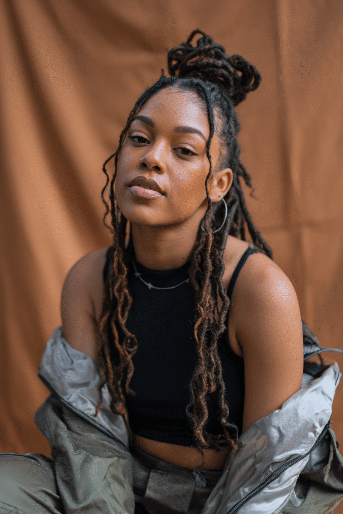A vibrant Black female model in her 20s with shoulder-to-waist-length loc wig, styled in a half-up loc twist with long locs flowing down, photographed against a seamless warm espresso backdrop with a satin finish. She has subtle makeup, a trendy outfit, a warm skin tone, and a friendly expression, captured with soft natural lighting.