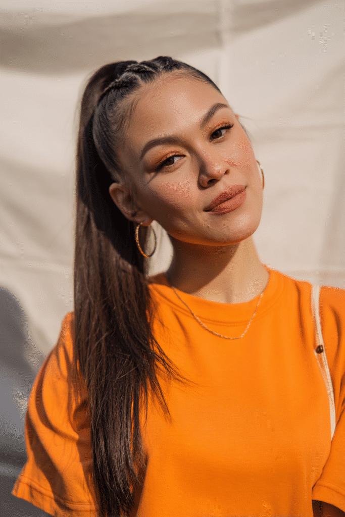 A vibrant female model in her 20s with shoulder-to-waist-length straight hair, styled in a low half-up Dutch braid with an inverted weave leading into a sleek ponytail down, photographed against a seamless light ivory backdrop with a velvety texture. She has subtle makeup, a trendy outfit, a warm skin tone, and a friendly expression, captured with soft natural lighting.