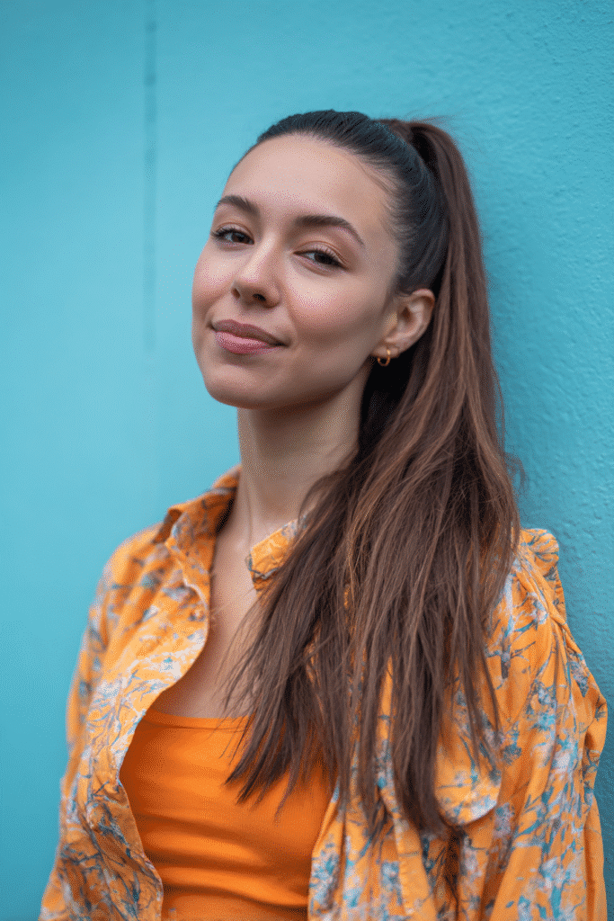 A vibrant female model in her 20s with shoulder-to-waist-length straight hair, styled in a low half-up side sweep with hair pulled to one side into a smooth ponytail down, photographed against a seamless soft aqua backdrop with a smooth surface. She has subtle makeup, a trendy outfit, a warm skin tone, and a friendly expression, captured with soft natural lighting.