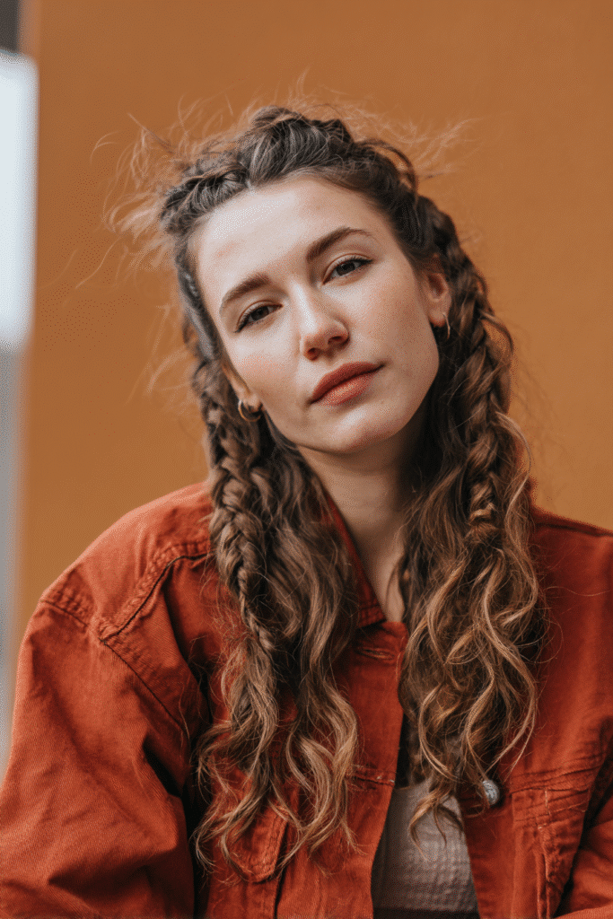 A vibrant female model in her 20s with shoulder-to-waist-length wavy hair, styled in a low half-up waterfall braid with cascading sections flowing into a loose ponytail down, photographed against a seamless warm saffron backdrop with a matte finish. She has subtle makeup, a trendy outfit, a warm skin tone, and a friendly expression, captured with soft natural lighting.