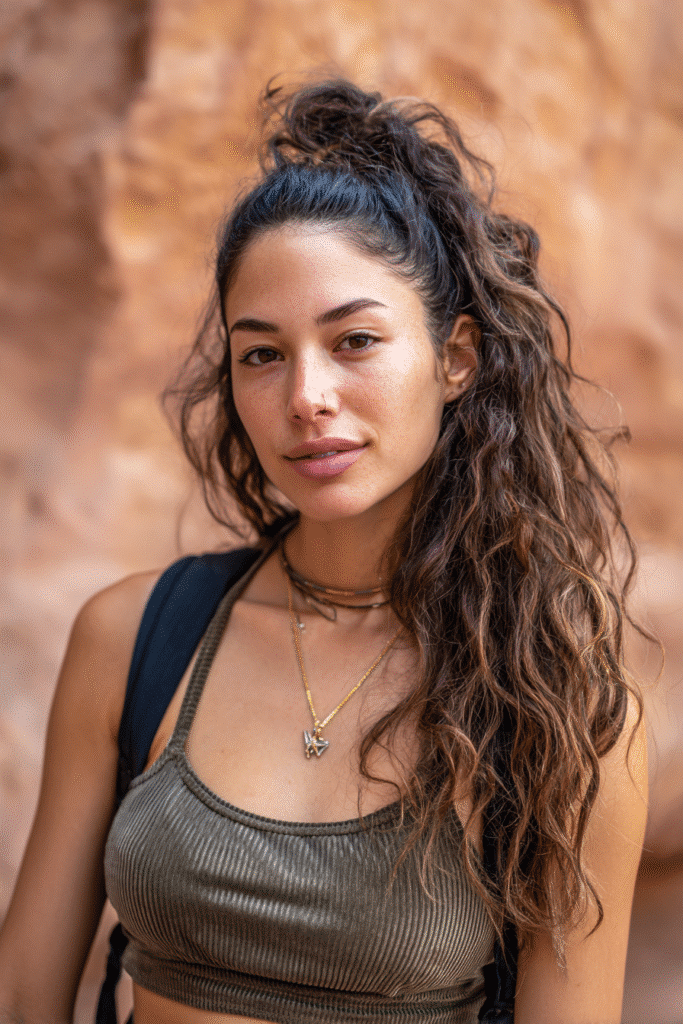 A vibrant female model in her 20s with shoulder-to-waist-length wavy hair, styled in a low half-up with boho vibes featuring textured strands into a loose ponytail down, photographed against a seamless warm sandstone backdrop with a velvety texture. She has subtle makeup, a trendy outfit, a warm skin tone, and a friendly expression, captured with soft natural lighting.