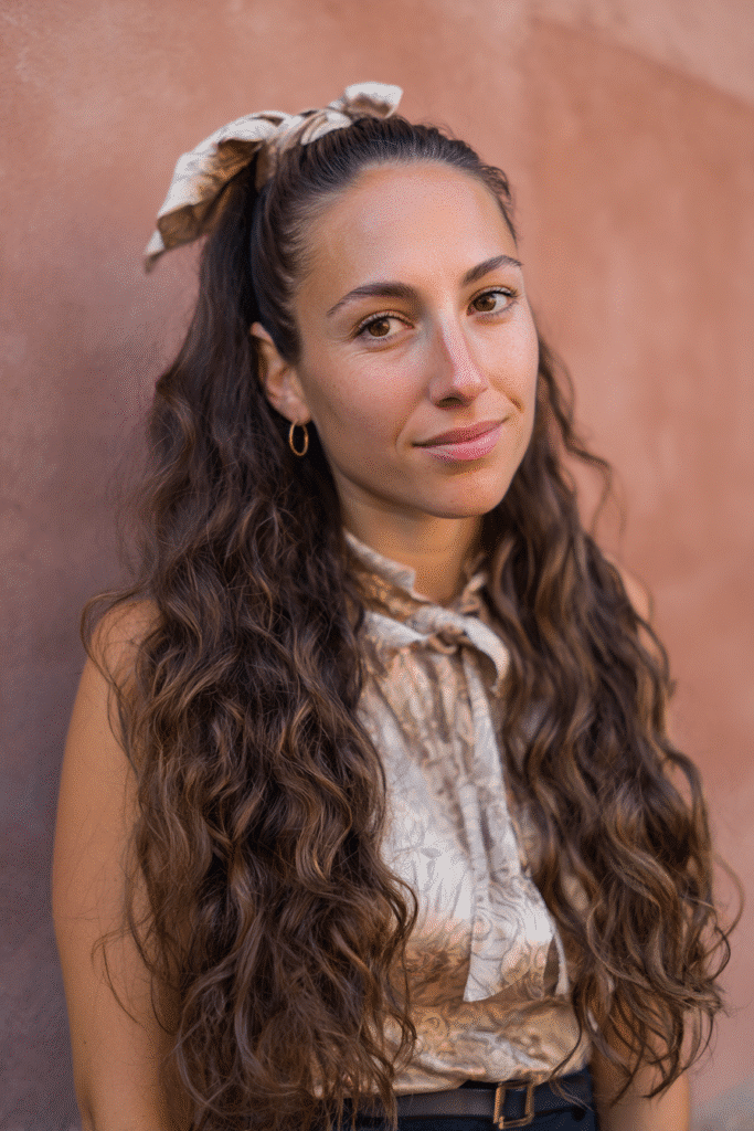 A vibrant female model in her 20s with shoulder-to-waist-length curly hair, styled in a low half-up secured with a ribbon tie, flowing into a bouncy ponytail down, photographed against a seamless warm rosewood backdrop with a satin finish. She has subtle makeup, a trendy outfit, a warm skin tone, and a friendly expression, captured with soft natural lighting.