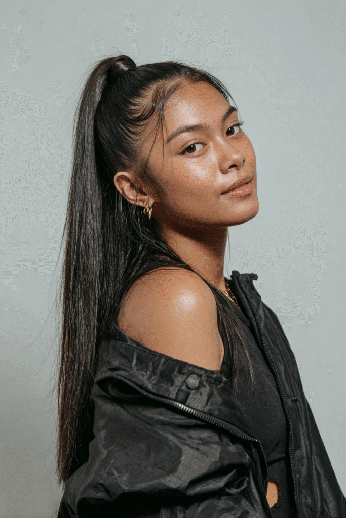 A vibrant female model in her 20s with shoulder-to-waist-length straight hair, styled in a low half-up with a volume crown, flowing into a sleek ponytail down, photographed against a seamless light indigo backdrop with a matte finish. She wears subtle makeup, a trendy outfit, and has a warm skin tone with a friendly expression, captured with soft natural lighting.