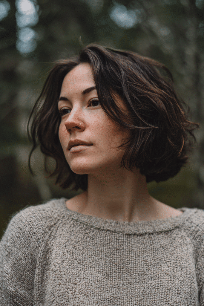 Casual American woman with thin hair styled in a messy low-maintenance French bob, wearing a knit sweater, captured in a natural forest scene.
