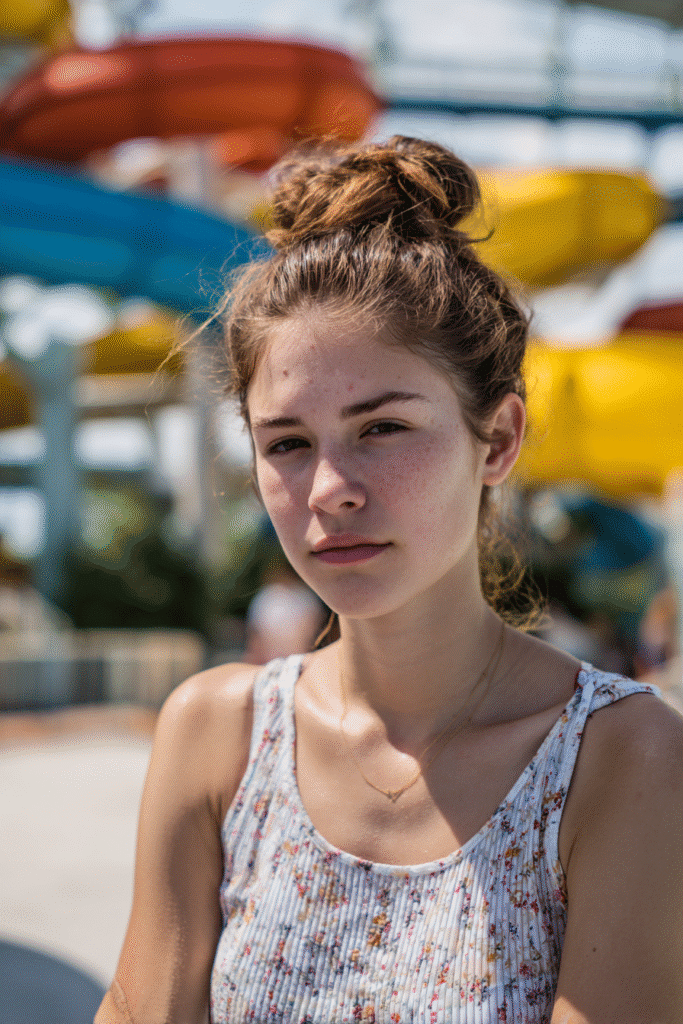 Smiling American teenage girl in a short-sleeve swim blouse with a neat low twisted bun, water slides visible in the background.