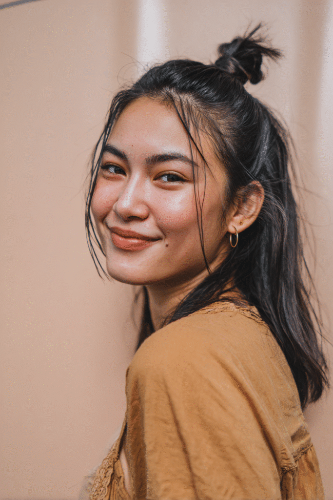A vibrant female model in her 20s with shoulder-to-waist-length straight hair, styled in a low twisted half-up bun flowing into a sleek ponytail down, photographed against a seamless soft blush backdrop with a silky finish. She wears subtle makeup, a trendy outfit, and has a warm skin tone with a friendly expression, captured with soft natural lighting.
