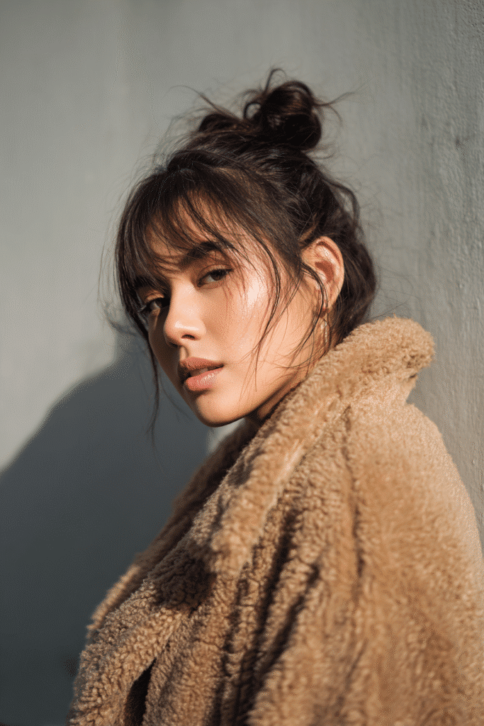 A vibrant female model in her 20s with shoulder-to-waist-length hair, styled in a messy bun with a casual chic look, framed from chest-level upward, photographed against a seamless light gray backdrop with a smooth surface. She has subtle makeup, a stylish winter coat, a warm skin tone, and a confident expression, captured with cozy winter lighting and a frosty glow.