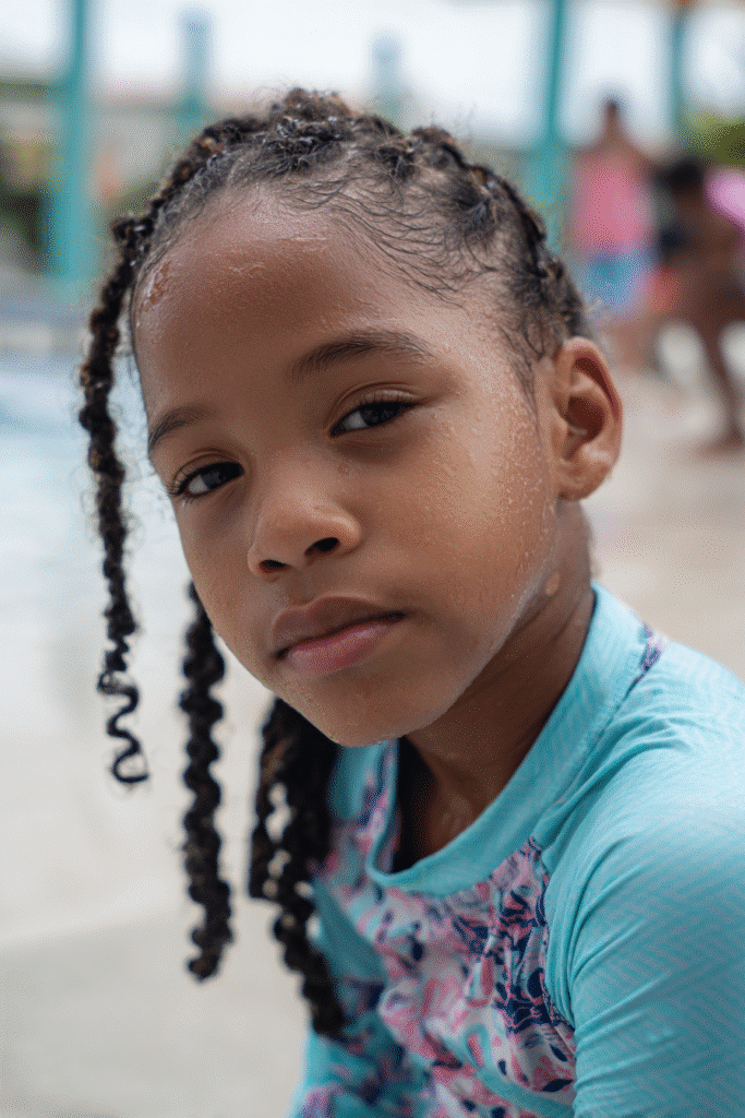 American girl styled with mini box braids for durability, modest aqua rash guard swim shirt, posing poolside with splashing kids.