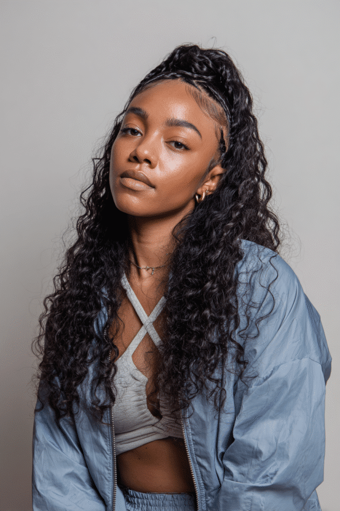 A vibrant Black female model in her 20s with shoulder-to-waist-length natural curl wig with clip-ins, styled in a half-up braided crown with natural curls flowing down, photographed against a seamless light mocha backdrop with a glossy sheen. She wears subtle makeup, a trendy outfit, and has a warm skin tone with a friendly expression, captured with soft natural lighting.