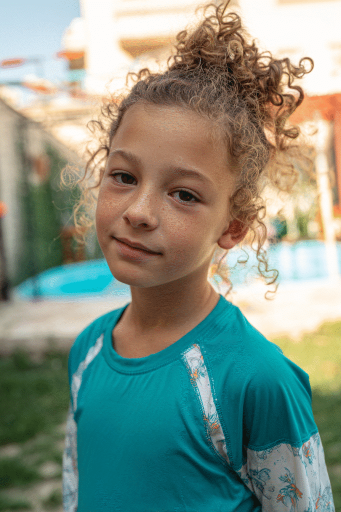 American girl with curly hair styled in a pineapple updo, wearing a full swim shirt, standing in a vibrant splash zone.