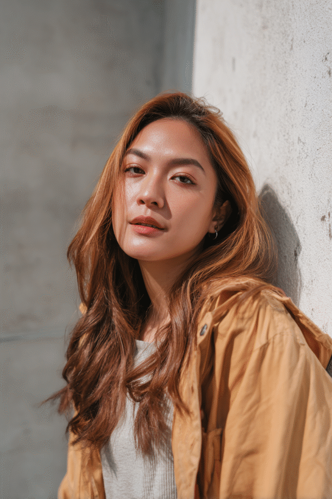 A vibrant American female model in her 20s with blended pumpkin spice balayage hair color, styled in a smooth hairstyle, photographed against a textured plaster backdrop with minimal texture. She wears subtle makeup, a trendy outfit, and has a warm skin tone with a friendly expression, captured with soft natural lighting.