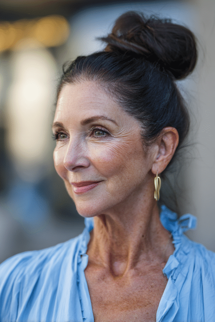 Realistic close-up of a woman over 60 with a dark brunette sleek top knot, minimal makeup, wearing a high-neck knit sweater, background of a muted beige wall.