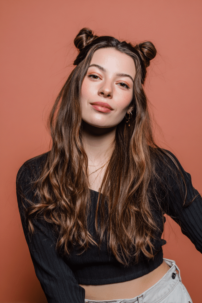 A vibrant female model in her 20s with shoulder-to-waist-length straight half wig, styled with space buns and loose strands flowing down, photographed against a seamless warm rose backdrop with a matte finish. She wears subtle makeup, a trendy outfit, and has a warm skin tone with a friendly expression, captured with soft natural lighting.