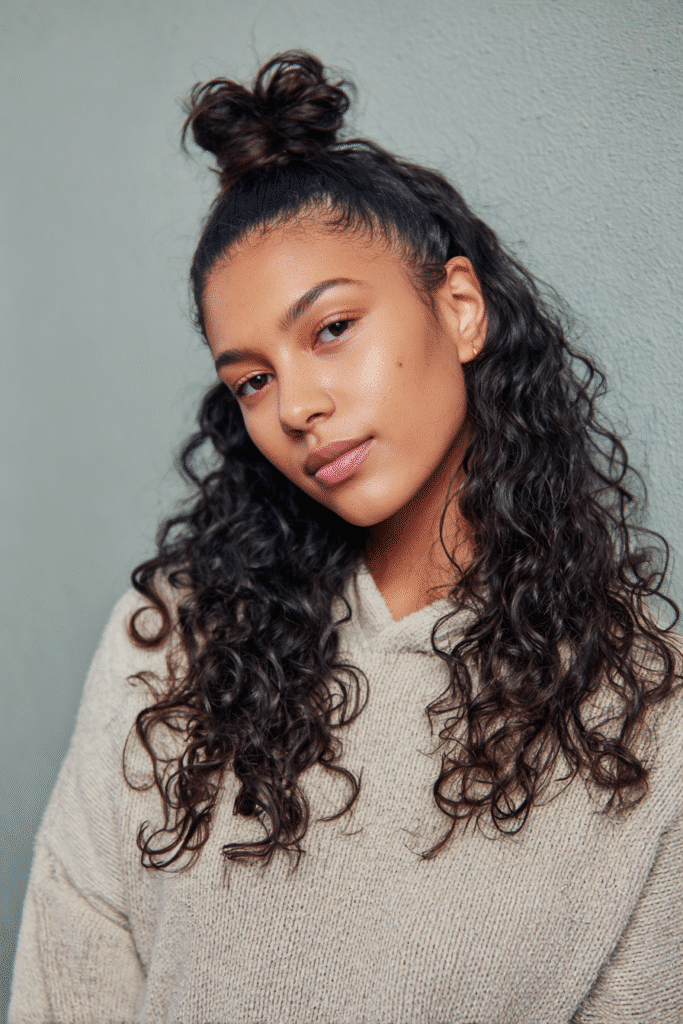 A vibrant female model in her 20s with shoulder-to-waist-length curly half wig, styled in a top knot with loose curls flowing down, photographed against a seamless light sage backdrop with a velvety texture. She has subtle makeup, a trendy outfit, a warm skin tone, and a friendly expression, captured with soft natural lighting.
