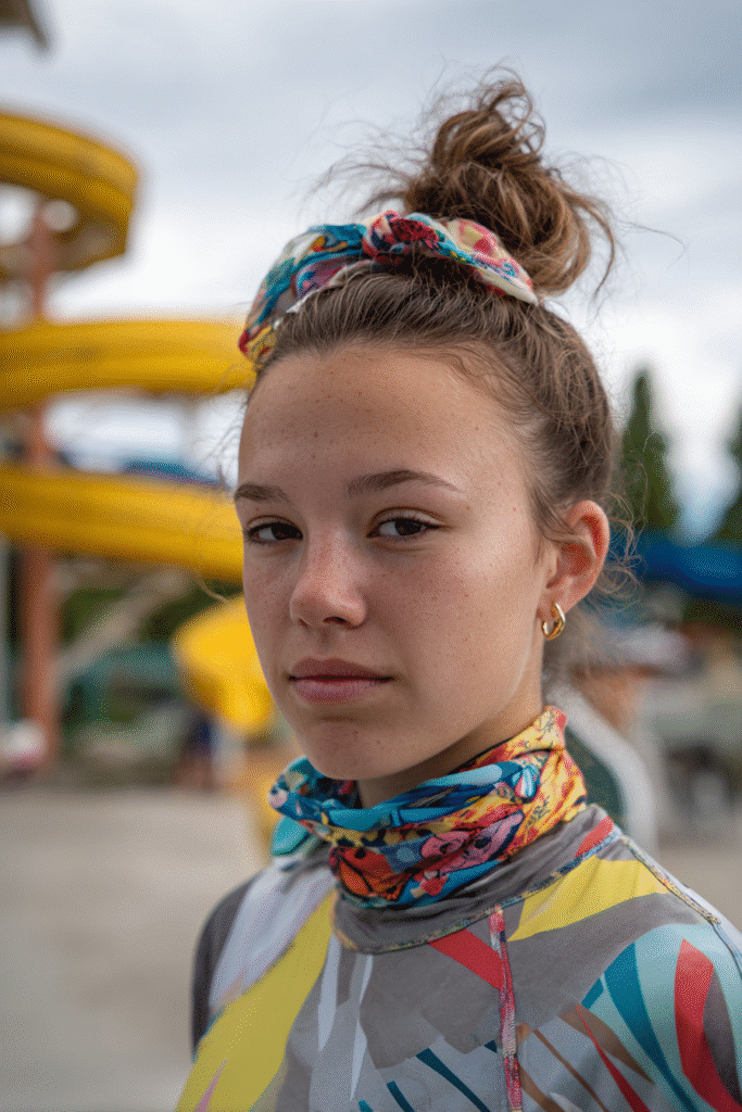 American teenage girl wearing a high top knot tied with a colorful scarf, modest patterned swim shirt, smiling near water slides.