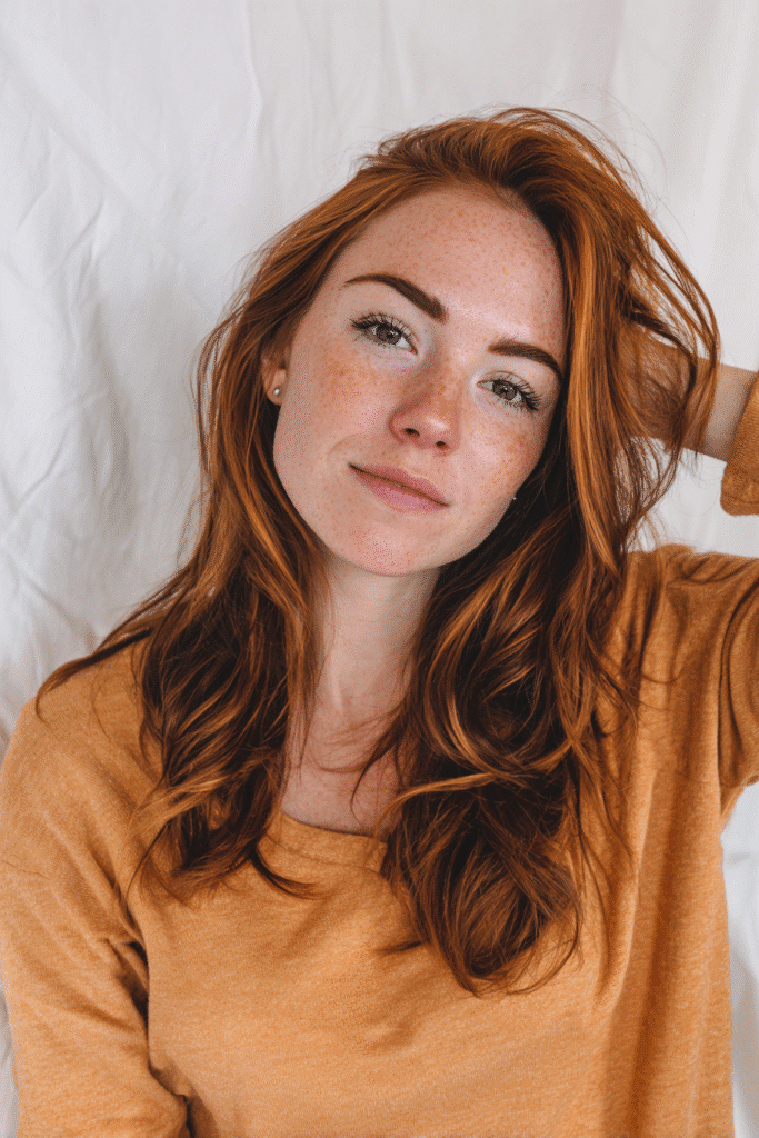 A vibrant American female model in her 20s with two-tone pumpkin spice hair color, styled in a modern hairstyle, photographed against a seamless white backdrop with minimal texture. She has subtle makeup, a trendy outfit, a warm skin tone, and a friendly expression, captured with soft natural lighting.