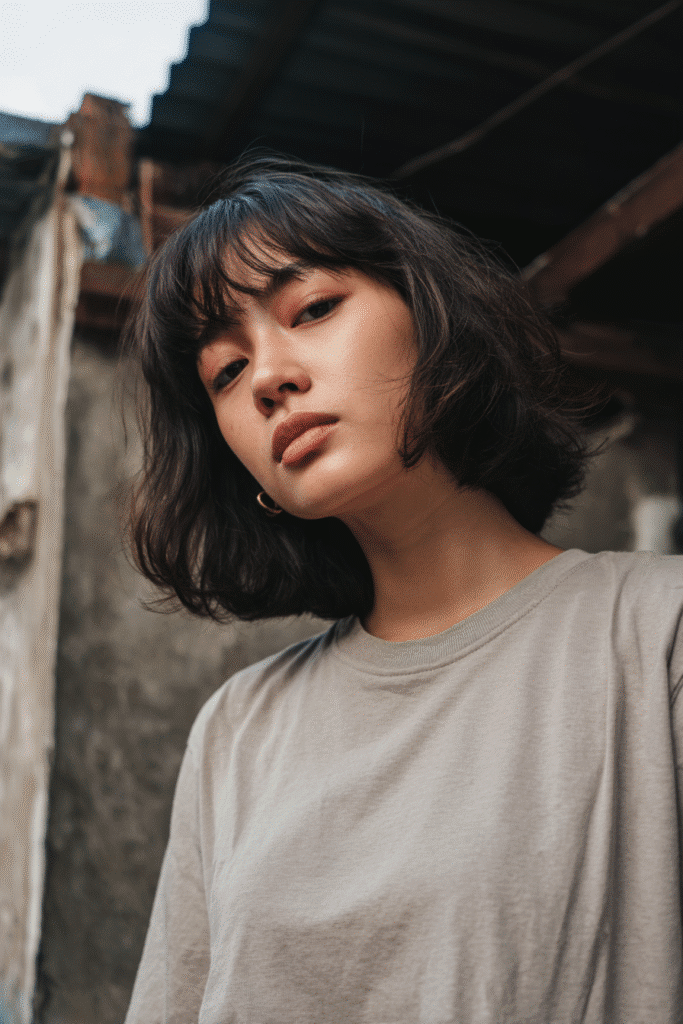 Trendy American model with thin hair in a two-toned French bob and choppy fringe, dressed in a modest full-sleeve shirt, rustic background.