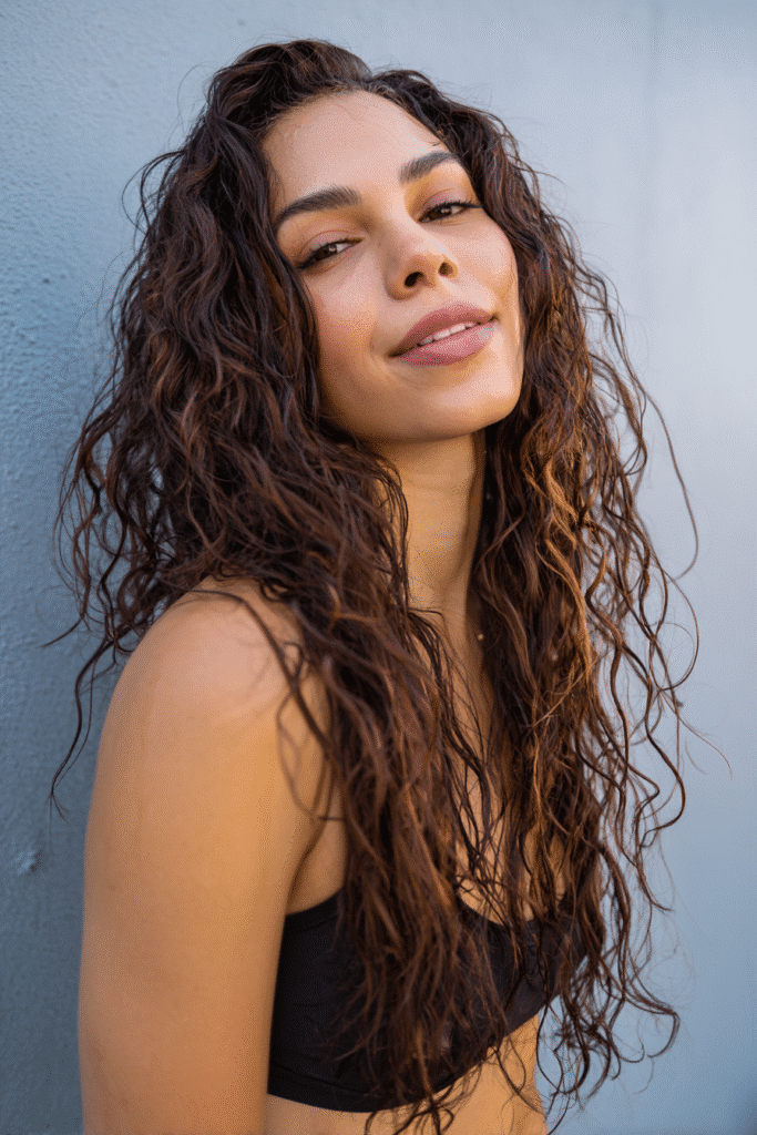 A vibrant female model in her 20s with shoulder-to-waist-length curly half wig, styled with a wet look and sleek, glossy curls flowing down, photographed against a seamless light indigo backdrop with a smooth surface. She has subtle makeup, a trendy outfit, a warm skin tone, and a friendly expression, captured with soft natural lighting.