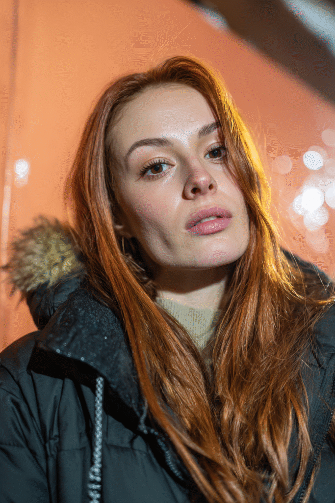A vibrant female model in her 20s with shoulder-to-waist-length wig hair, styled in a sleek protective wig with a natural finish, captured in a close-up shot focusing on the hair, photographed against a seamless warm peach backdrop with a glossy sheen. She has subtle makeup, a stylish winter coat, a warm skin tone, and a confident expression, captured with cozy winter lighting and a frosty glow.