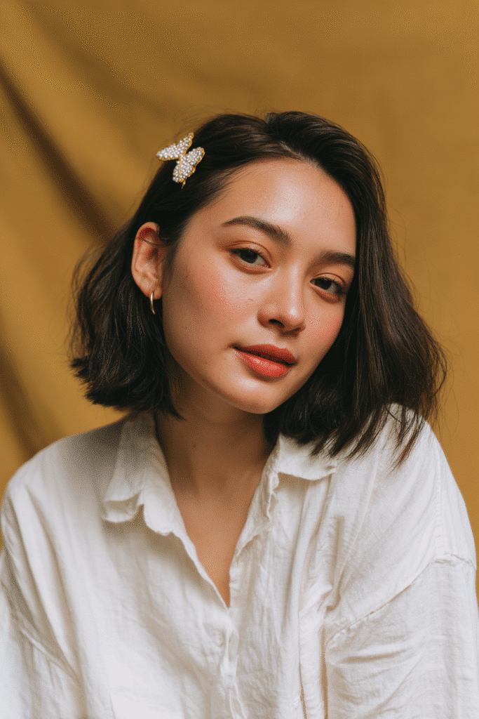 A gorgeous young female model in her 20s with natural skin tone, showcasing a prom hairstyle for short hair with an angled bob and statement clip, featuring a decorative clip for accent, captured in a close-up shot focusing on the hair, photographed against a seamless warm gold backdrop with glossy sheen. She has subtle makeup, an elegant full-shirt prom dress, realistic hair texture, warm skin tone, and a friendly expression, captured with soft natural lighting and an Instagram-style beauty vibe.