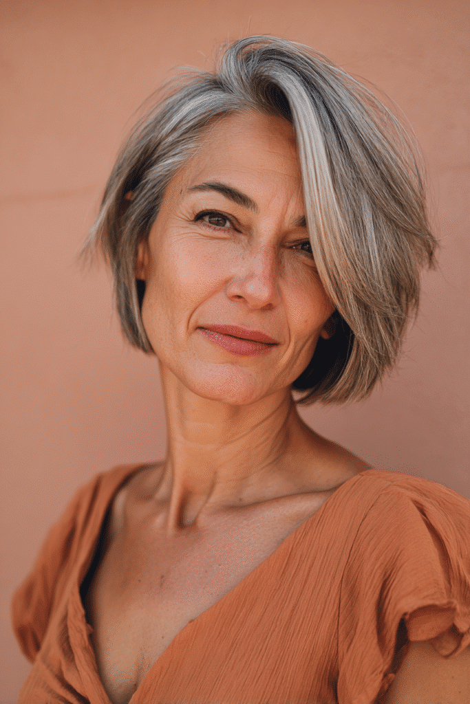 A vibrant American female model over 50 with a mature appearance, showcasing a youthful Asymmetrical Bob hairstyle with uneven lengths for a modern edge, captured in a close-up shot focusing on the hair, photographed against a seamless warm peach backdrop with a matte finish. She has subtle makeup with visible aging, an elegant outfit, realistic hair texture with silver highlights, warm skin tone, and a friendly expression, captured with soft natural lighting and an Instagram-style beauty vibe