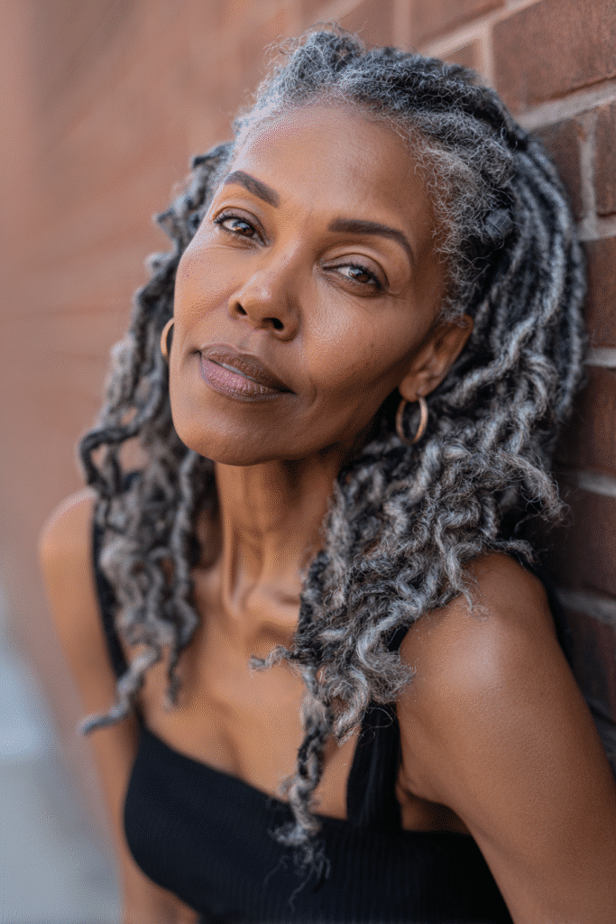 An elegant Black woman over 50 with a warm skin tone and friendly expression, showcasing bold two-tone twists braids hairstyle for older Black women over 50. The hair is two-tone black and silver, set against an urban brick wall backdrop with minimal texture. She wears subtle makeup and an elegant outfit suitable for mature women, captured in soft natural lighting for an Instagram-style beauty photo.