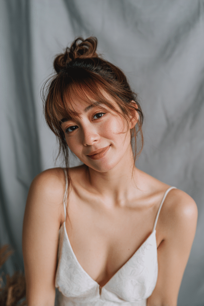 A gorgeous young American bride with a warm skin tone and friendly expression, showcasing bottleneck bangs with low bun wedding hairstyle. The hair is an elegant low bun with bottleneck-shaped bangs, colored caramel brown, styled elegantly. She is dressed in an elegant bridal gown, captured in soft natural lighting with subtle makeup, set against a soft gray fabric backdrop with minimal texture for an Instagram-style beauty photo.