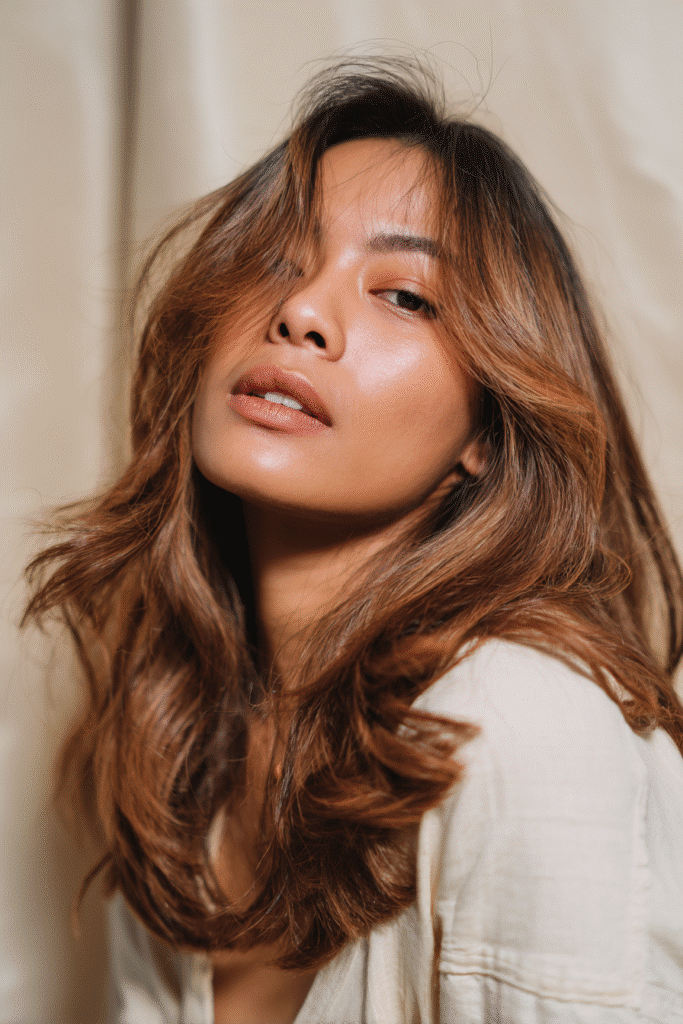 A vibrant female model in her 20s with shoulder-to-waist-length hair, styled in a caramel brunette balayage with soft highlights, captured in a close-up shot focusing on the hair, photographed against a seamless soft ivory backdrop with a satin finish. She has subtle makeup, a trendy outfit, a warm skin tone, and a confident expression, captured with warm natural lighting and a soft glow.