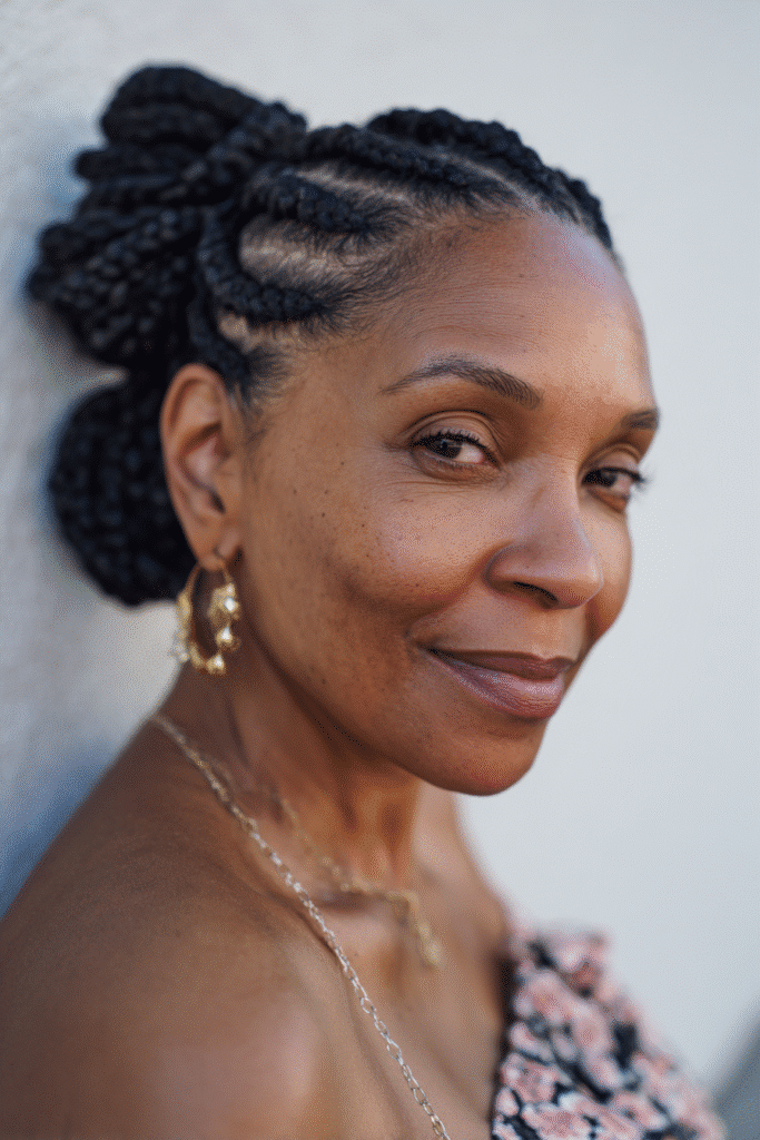 An elegant Black woman over 50 with a warm skin tone and friendly expression, showcasing classic black box braids hairstyle for older Black women over 50. The hair is black, set against a seamless white backdrop with minimal texture. She wears subtle makeup and an elegant outfit suitable for mature women, captured in soft natural lighting for an Instagram-style beauty photo.