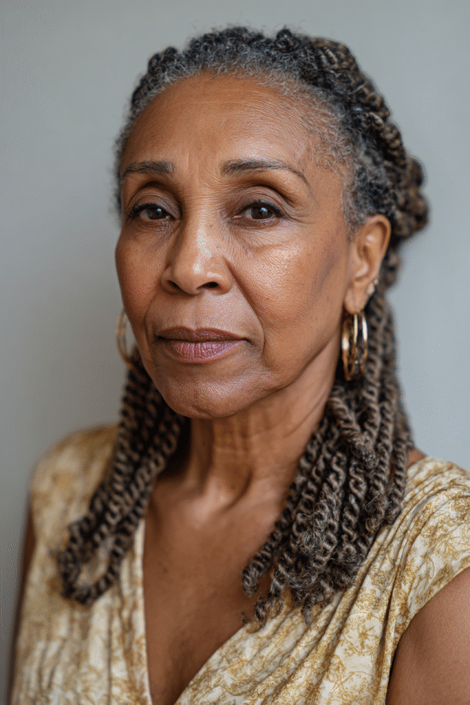 An elegant Black woman over 50 with a warm skin tone and friendly expression, showcasing classic passion twists braids hairstyle for older Black women over 50. The hair is natural black, set against a textured plaster backdrop with minimal texture. She wears subtle makeup and an elegant outfit suitable for mature women, captured in soft natural lighting for an Instagram-style beauty photo.